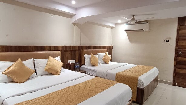 Habitación de hotel con dos camas, cada una con cabecero de madera, ropa de cama blanca y almohadones color beige. Se observa un ventilador de techo, un equipo de aire acondicionado y una botella de agua sobre la mesita de noche.