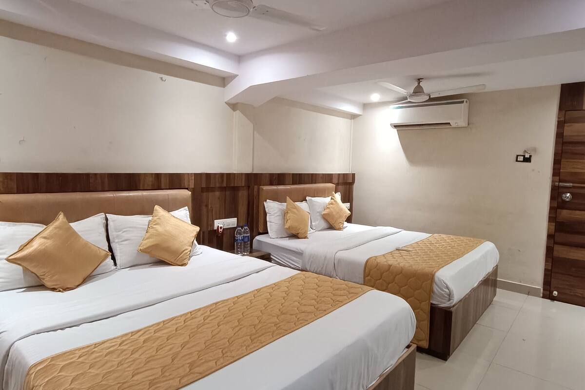 A hotel room with two beds, each with a wooden headboard, white bedding, and beige throw pillows. There is a ceiling fan, an air conditioning unit, and a water bottle on the nightstand.