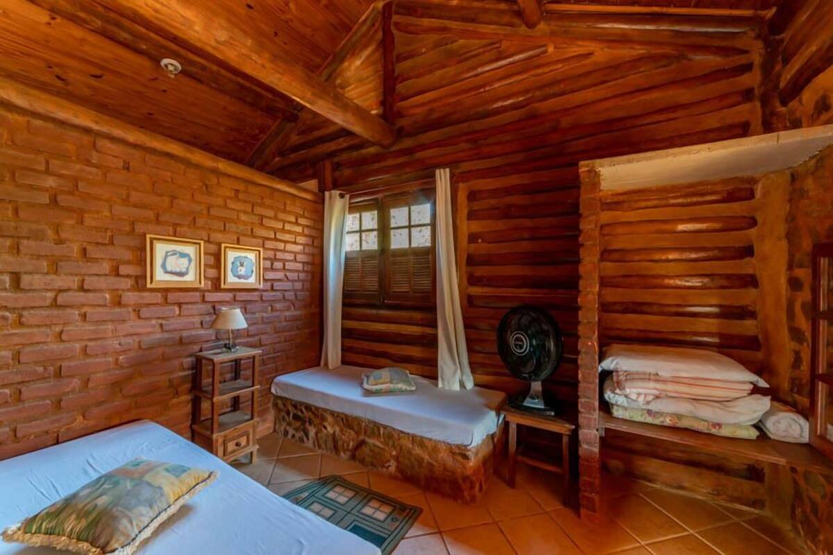 A rustic cabin room with a wooden ceiling, a brick wall, a bed, a bench, a fan, and a window with curtains.