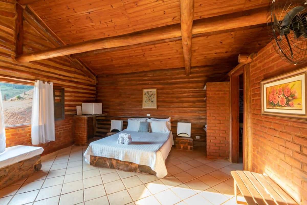 A wooden cabin bedroom with a bed, bedside tables, a bench, and a window with curtains.