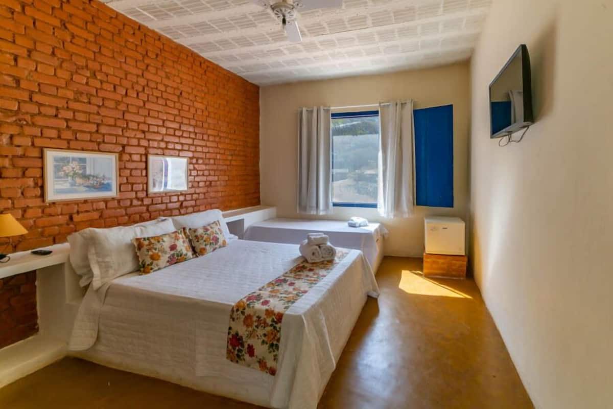 A hotel room with a brick wall, a bed with a floral bedspread, a bedside table with a lamp, a window with curtains, and a television mounted on the wall.