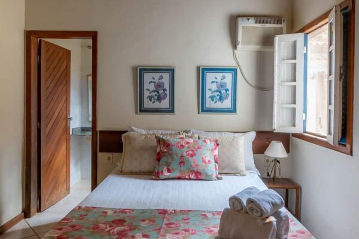 A bedroom with a wooden ceiling, a bed with floral bedding, two framed pictures on the wall, and a window with open shutters.