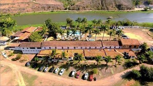 A large, rectangular building with a flat roof, surrounded by a parking lot with several cars, palm trees, and a body of water in the background. - Hotel Fazenda Santa Helena (Simao Pereira)