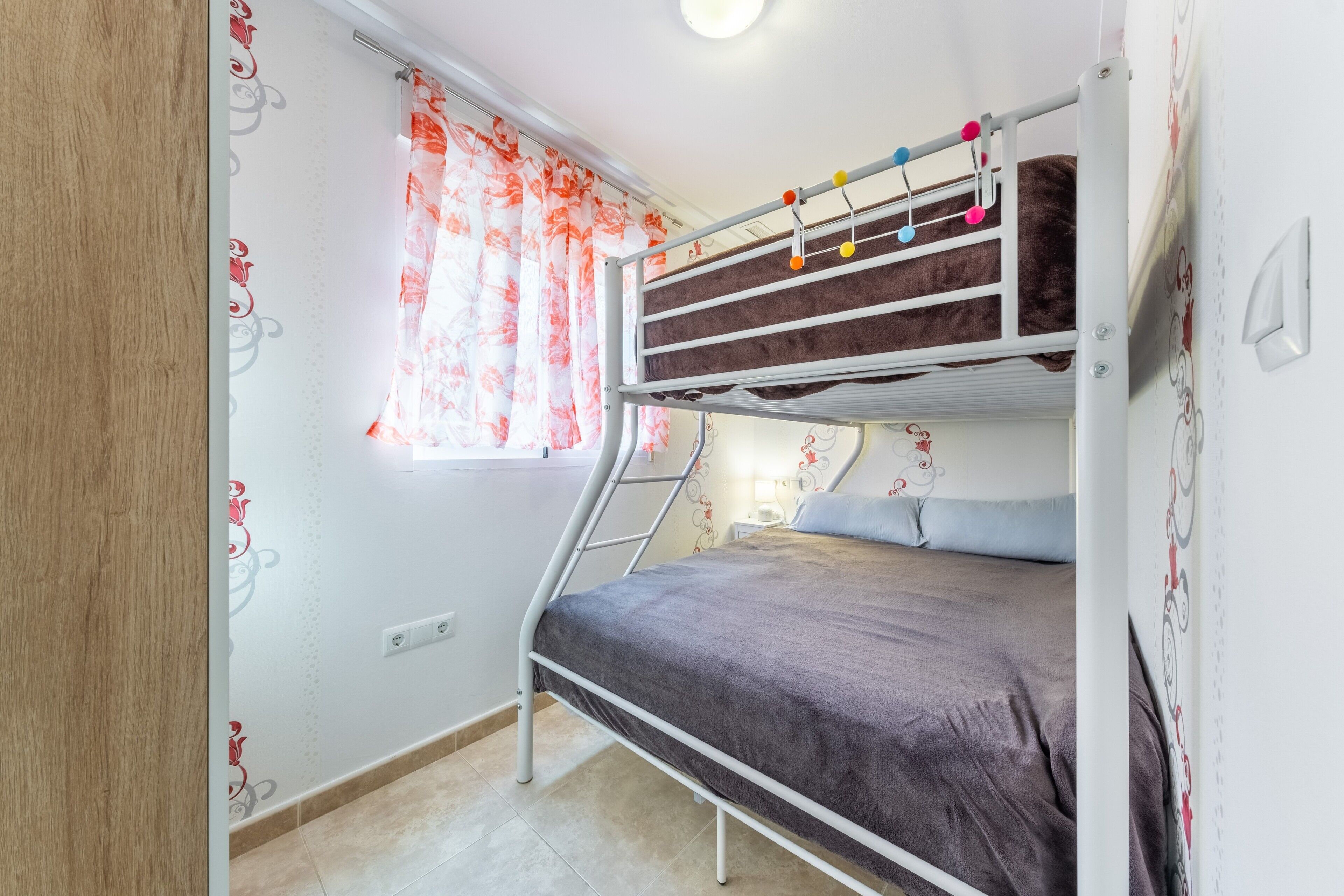 Een stapelbed met raam en gordijnen, een muur met bloemenbehang, en een lichtschakelaar.