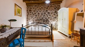 Une chambre avec un mur en pierre, un lit avec un cadre en métal, un bureau en bois, une chaise, une armoire et un miroir.
