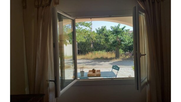 Una ventana abierta con cortinas, vista de follaje al exterior y una mesa con objetos encima.