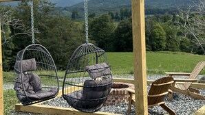 A wooden swing set with two hanging chairs, a brick fire pit, and wooden Adirondack chairs in a grassy outdoor area with a mountainous backdrop.