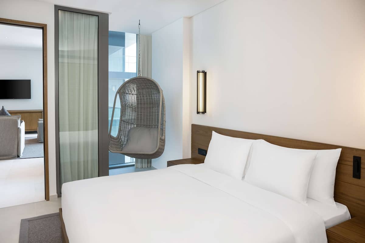 A modern hotel room with a large bed, a hanging chair, a TV, and a window with a view.