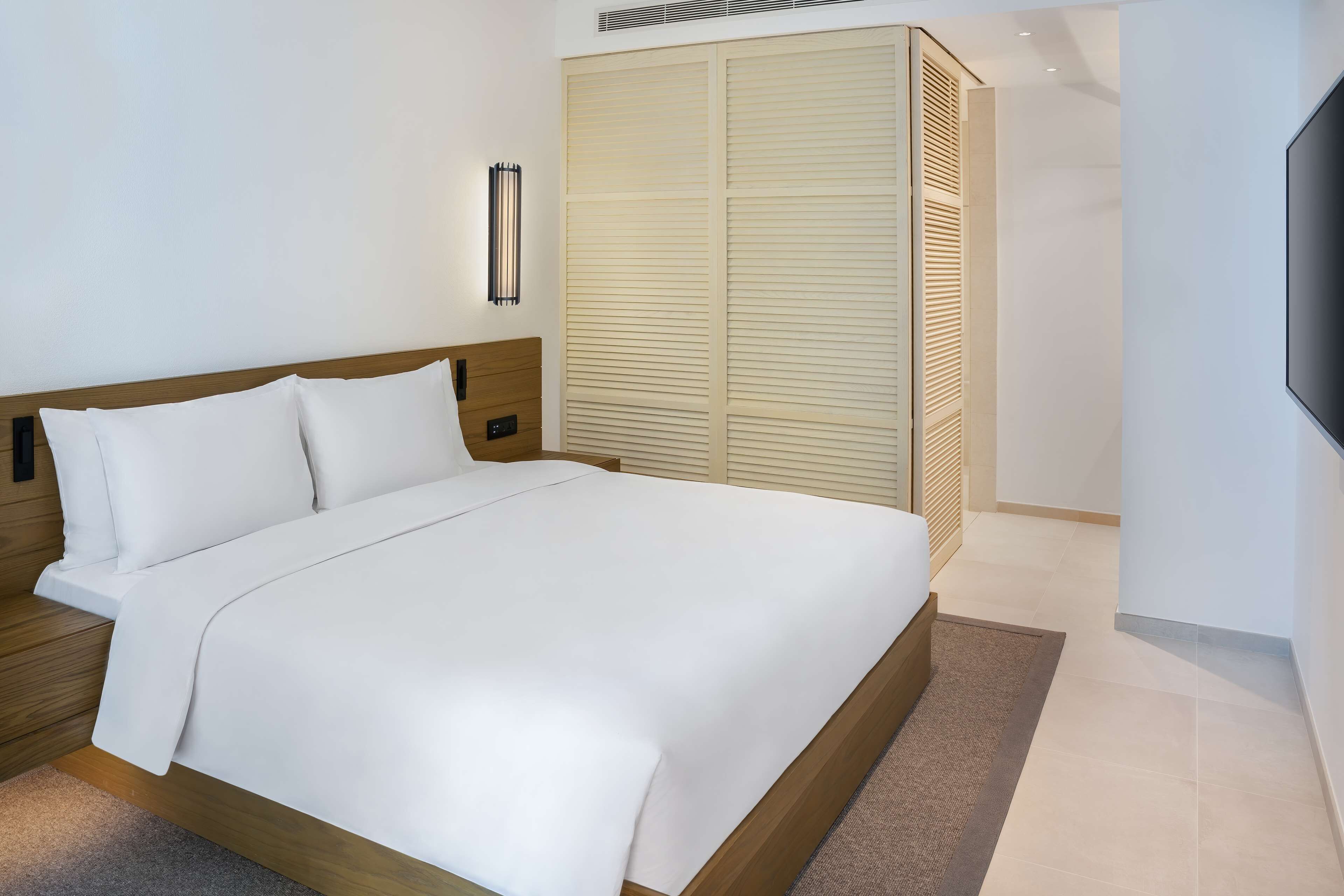 A modern hotel room with a large bed, wooden headboard, a built-in wardrobe, and a flat-screen TV mounted on the wall.