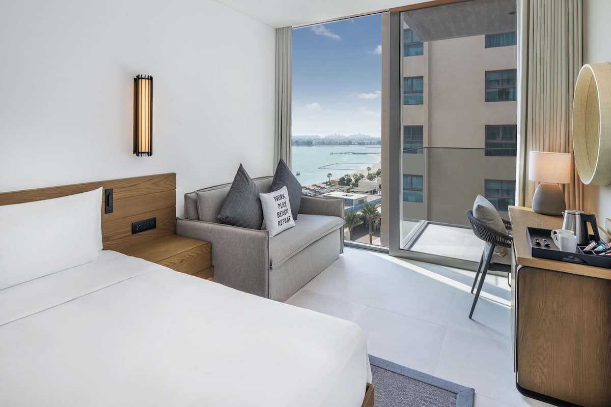 A modern hotel room with a large bed, a sofa with a city view, a desk with a lamp, and a large window overlooking a body of water.