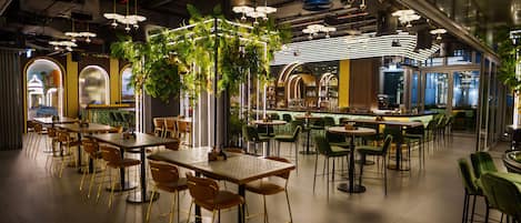 A spacious indoor dining area with multiple tables and chairs, a central plant installation, and a bar area in the background.