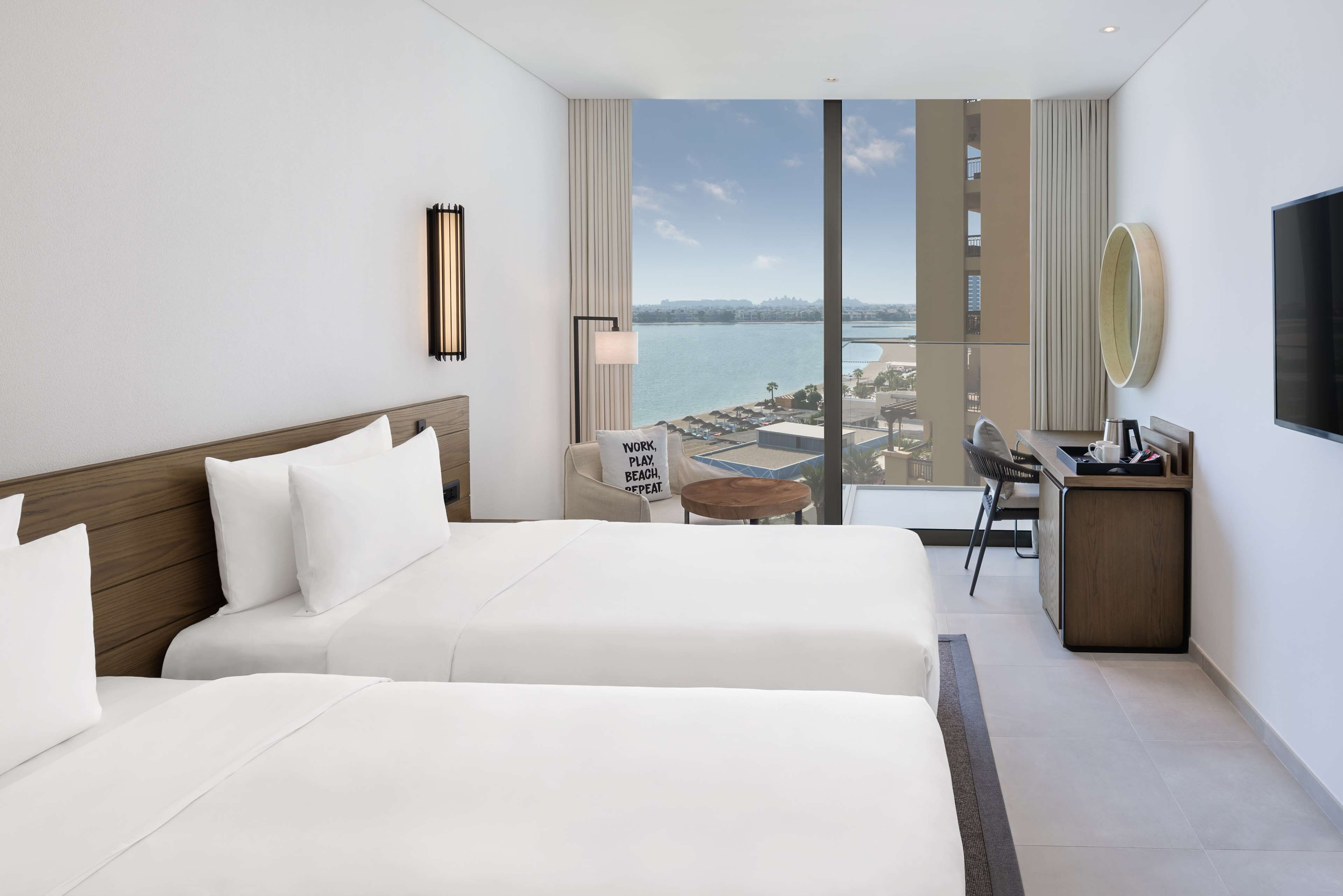 Ein Hotelzimmer mit zwei Betten, einem Schreibtisch, einem Sessel, einem kleinen Tisch, einem Fernseher und Blick auf das Meer.