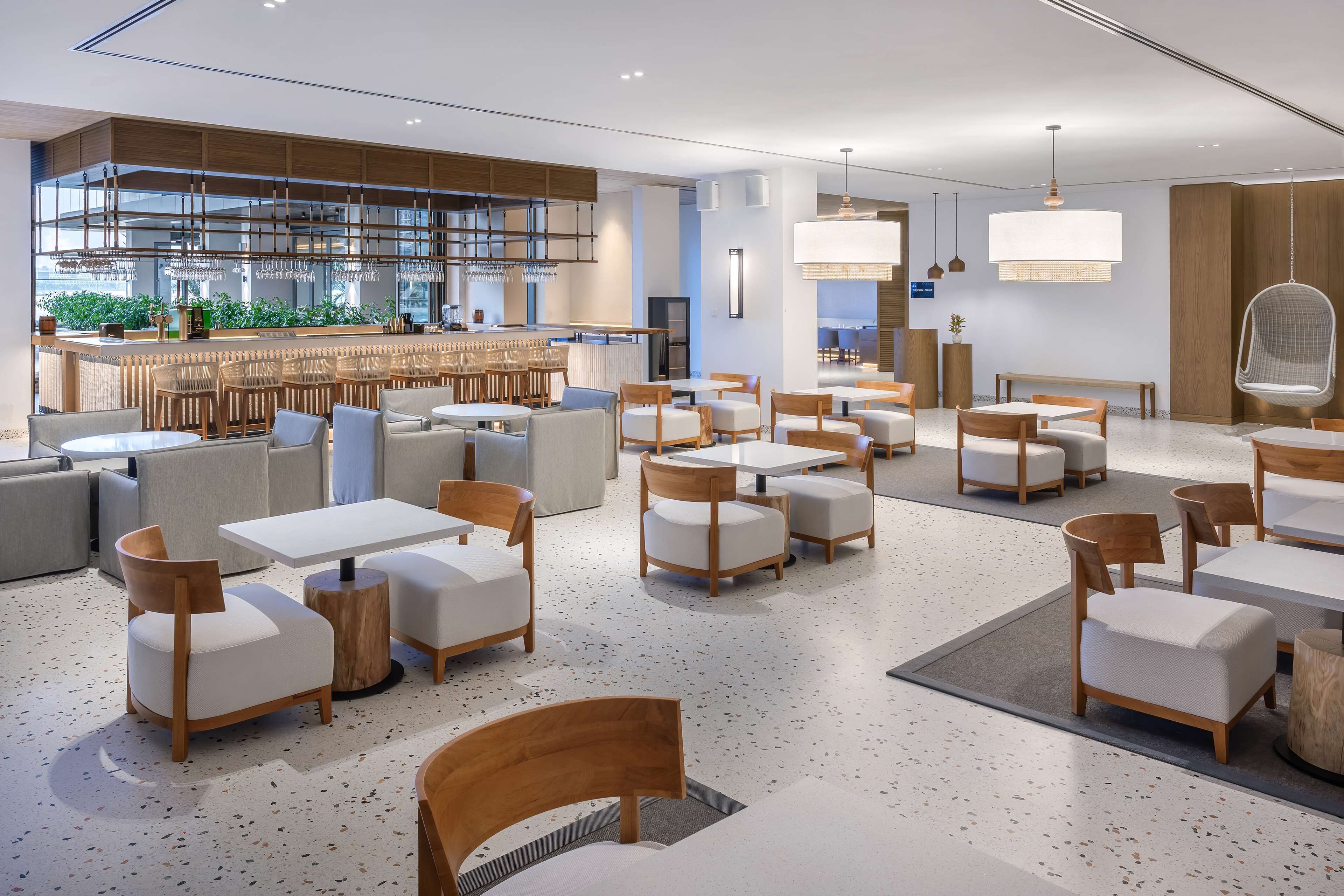 Un restaurante moderno con zona de bar, asientos con sillas y mesas de madera, y suelo con textura moteada.