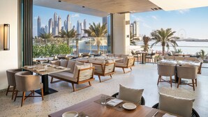 A spacious outdoor dining area with multiple seating arrangements, a view of a city skyline, and a body of water in the background. - Radisson Beach Resort Palm Jumeirah (Dubai)