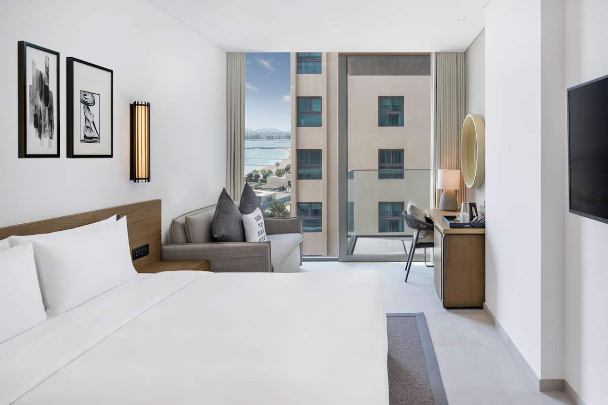 A hotel room with a large bed, a sofa, a desk, and a TV. There are framed pictures on the wall and a view of the city from the window.