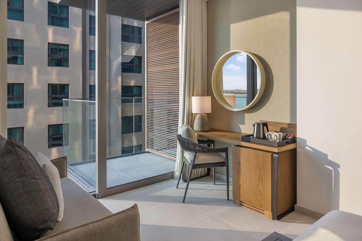 A modern hotel room with a balcony, a desk with a chair, a round mirror, and a view of the city.