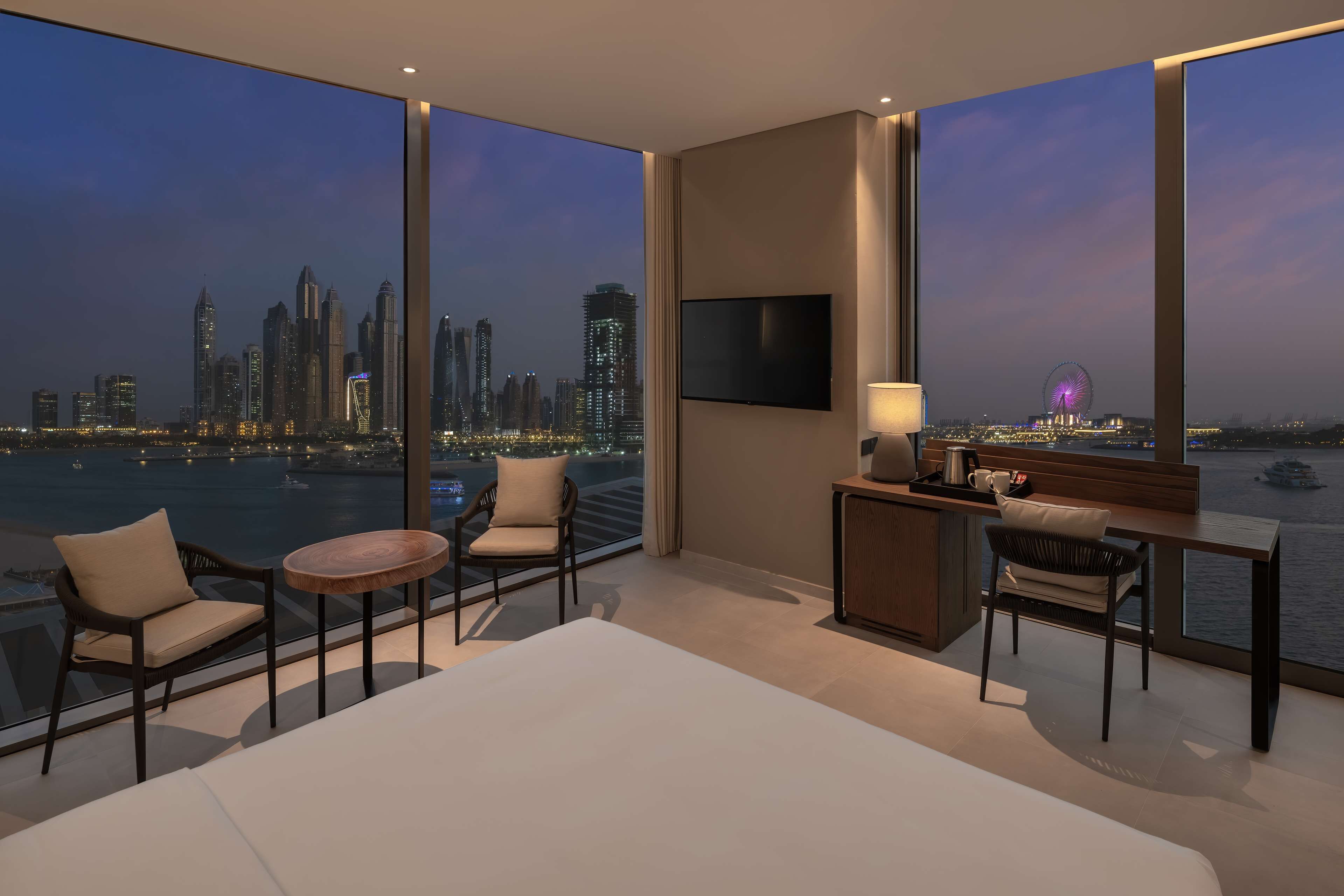 A hotel room with a large window offering a city skyline view, a bed, two chairs, a small table, and a TV mounted on the wall.
