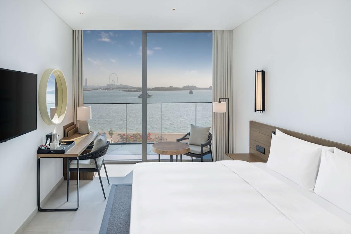 A hotel room with a large bed, a desk with a chair, a TV, and a view of a body of water with a bridge.