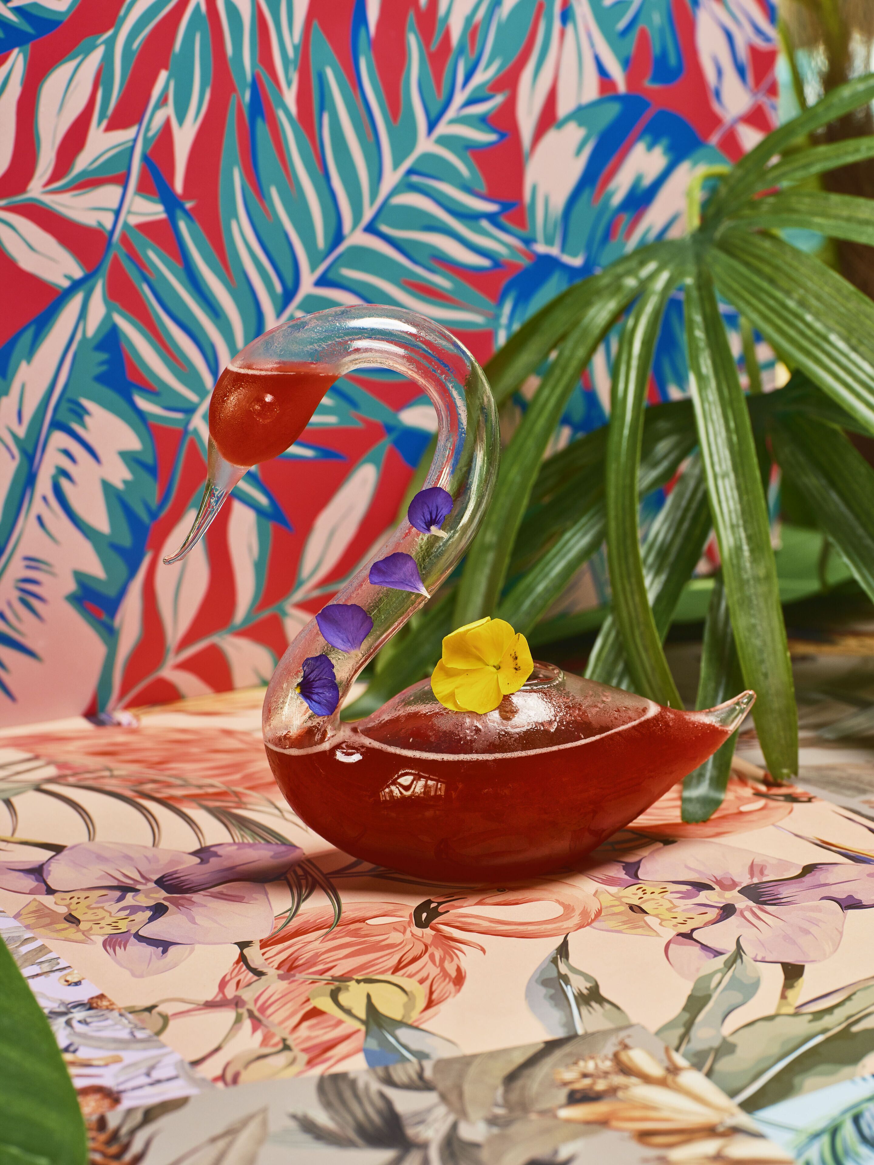 A decorative glass teapot with a swan design, placed on a floral-patterned tablecloth.