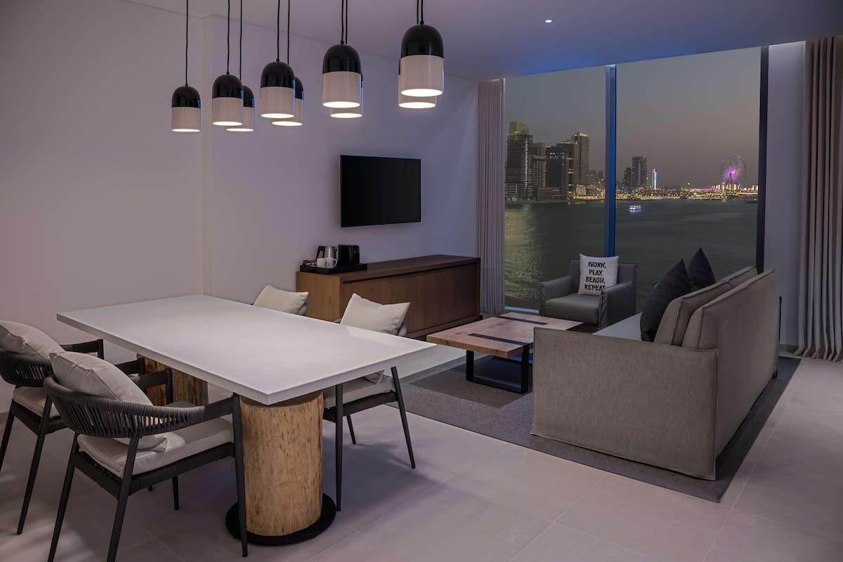 A modern hotel room with a dining area, a sofa, a TV, and a large window with a city view.