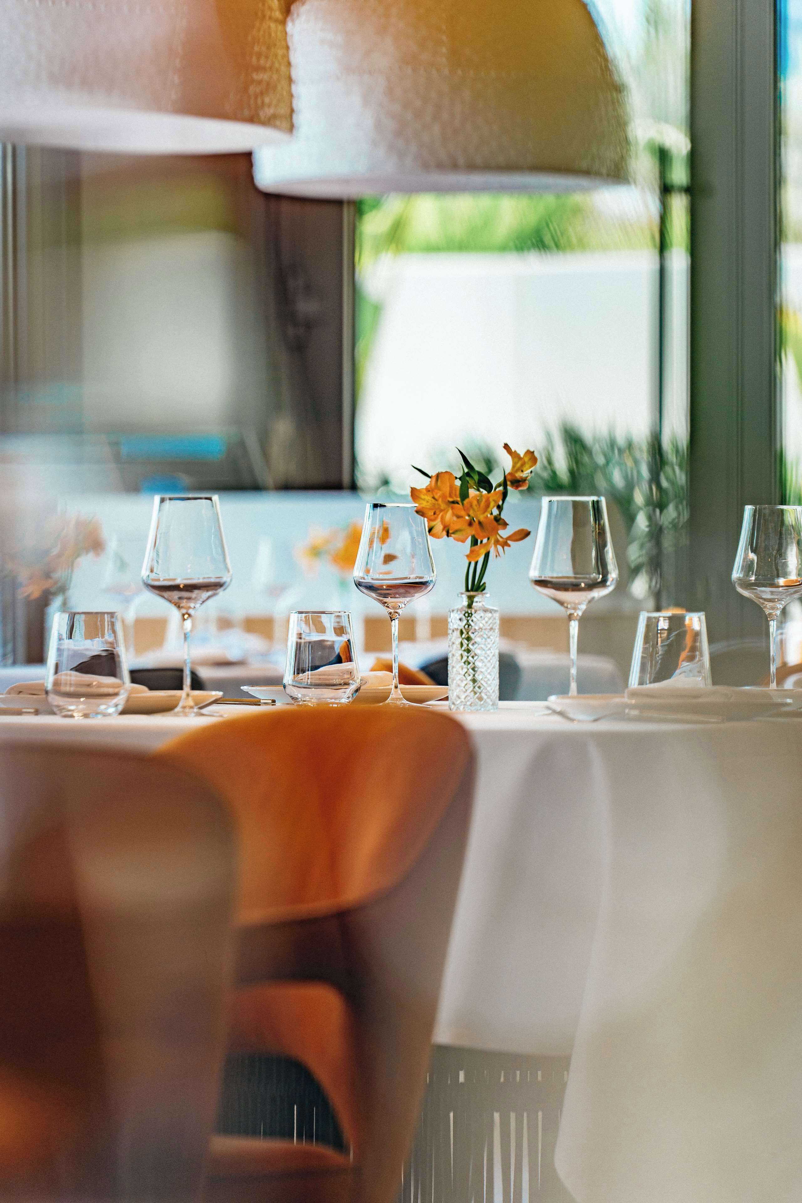 Une table à manger dressée avec des verres à vin et un vase rempli de fleurs, avec une fenêtre en arrière-plan.