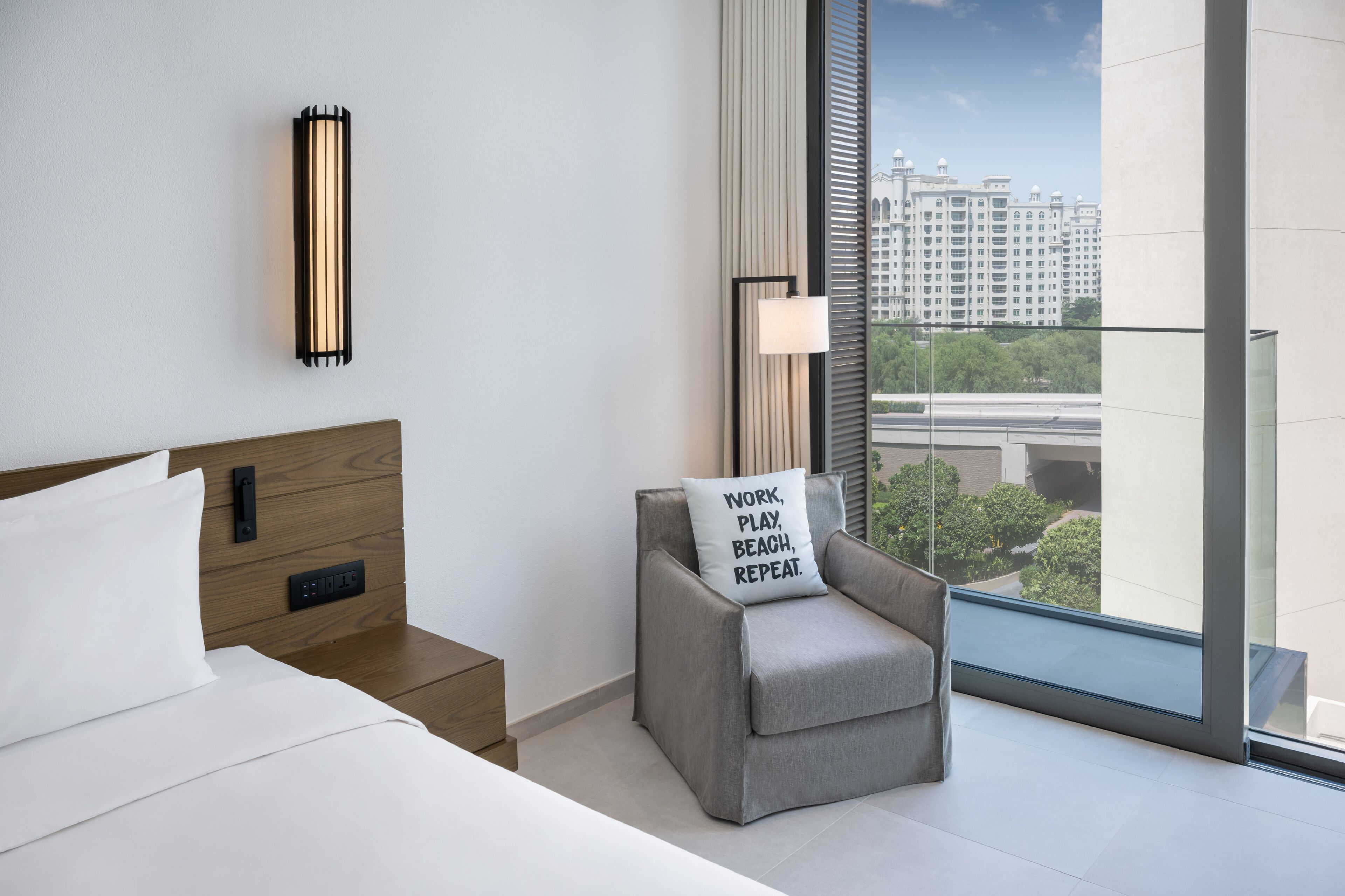 A modern hotel room with a large bed, a grey armchair with a motivational pillow, a bedside table with a lamp, and a view of a cityscape through floor-to-ceiling windows.