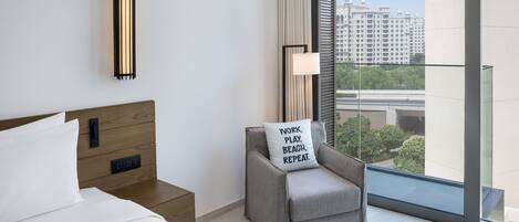 A modern hotel room with a large bed, a grey armchair with a motivational pillow, a bedside table with a lamp, and a view of a cityscape through floor-to-ceiling windows.
