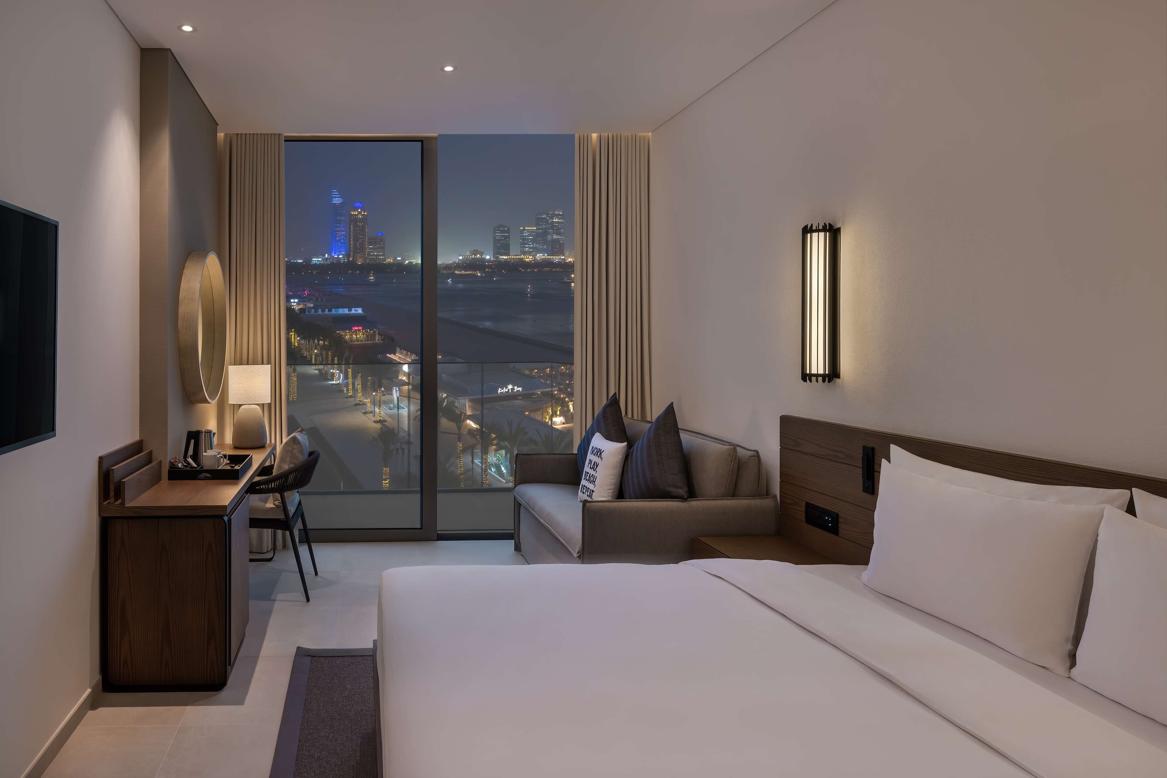 Quarto de hotel com uma cama grande, uma mesa com cadeira, um sofá e vista para a cidade à noite.