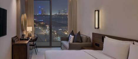 Quarto de hotel com uma cama grande, uma mesa com cadeira, um sofá e vista para a cidade à noite.
