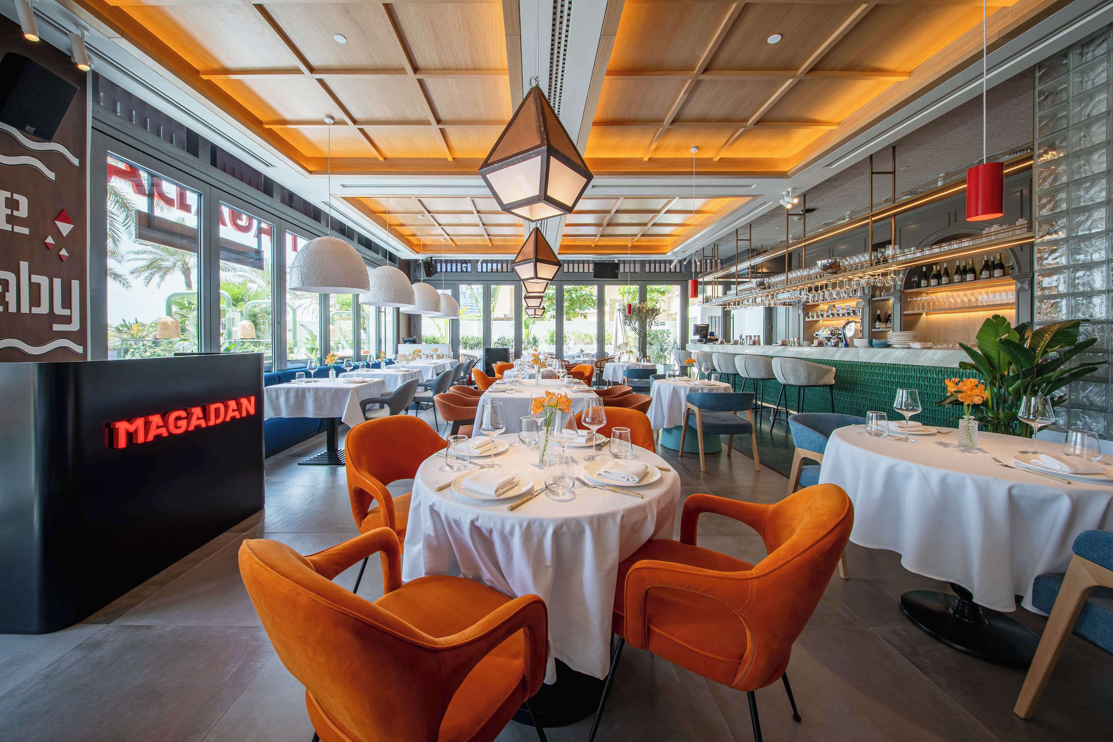 A restaurant with round tables set for dining, orange chairs, and a bar area with shelves of glasses and bottles.