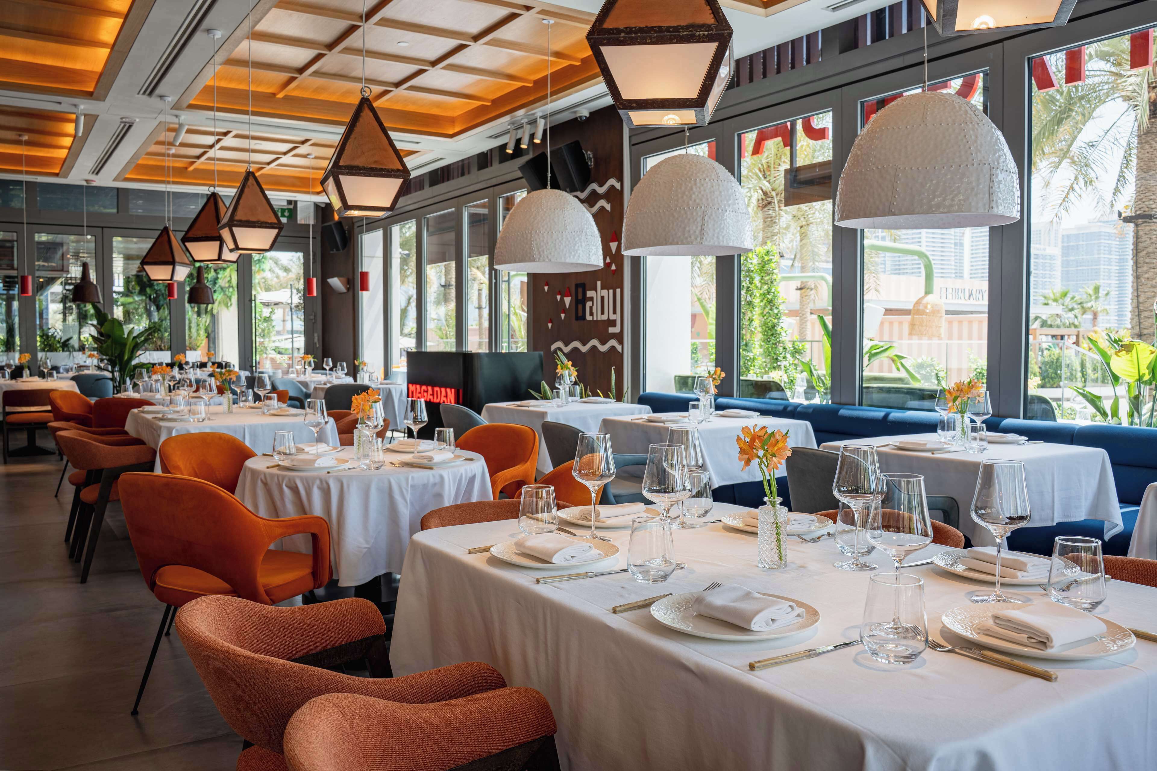 Ein Restaurant mit mehreren Tischen, gedeckt für das Essen, mit weißen Tischdecken und orangefarbenen Stühlen.