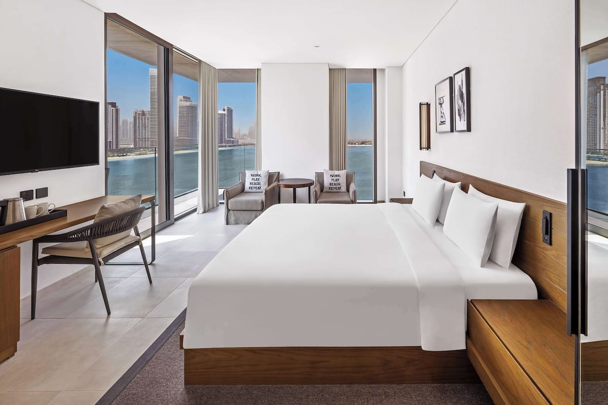 A modern hotel room with a large bed, a desk, a chair, a TV, and a balcony with a city view.