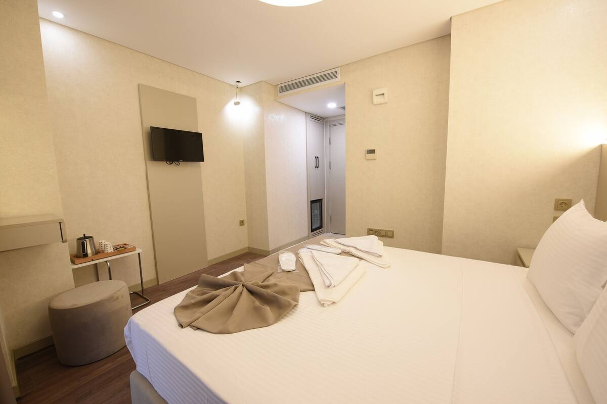 A hotel room with a bed, a TV mounted on the wall, a small table with a kettle, and a trash bin.