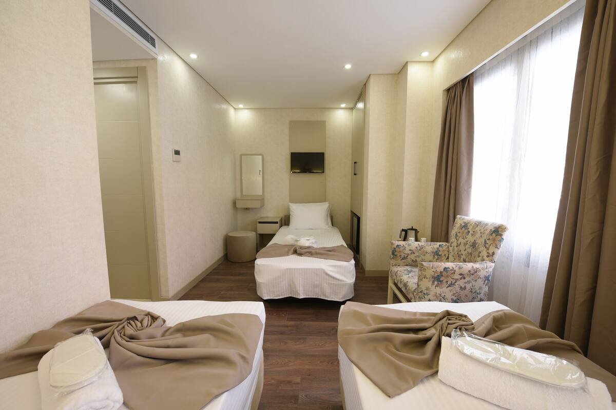 A hotel room with two beds, a sitting area with a chair, a TV mounted on the wall, and a window with curtains.