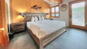 A bedroom with a wooden headboard, a bed with white bedding, a nightstand, a wall-mounted lamp, a window with blinds, and a door with a glass panel.