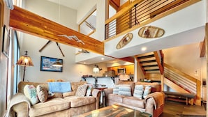 A spacious living area with a high ceiling, a wooden staircase, and a kitchen in the background.