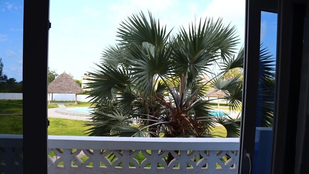 A view through a window with a white lattice fence, a palm tree, and a thatched-roof structure in the background. - Stay.Plus Kijani Apartments (Malindi)