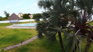 A pool surrounded by a grassy area with palm trees and a thatched-roof structure. - Stay.Plus Kijani Apartments (Malindi)