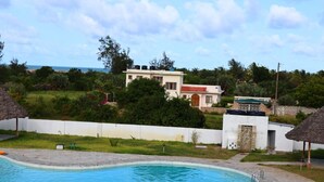 A swimming pool with a stone border, a white building with a red roof in the background, and a thatched-roof structure to the left. - Stay.Plus Kijani Apartments (Malindi)