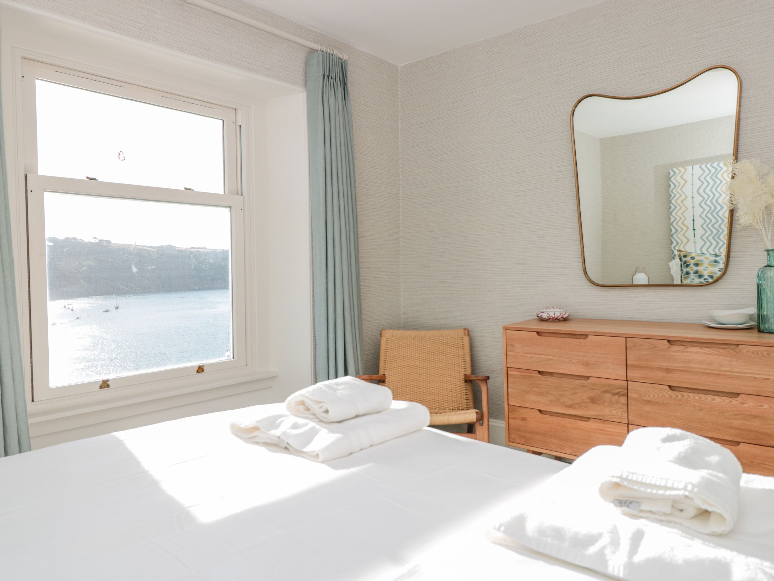 Une chambre d’hôtel avec un lit, une commode en bois, une chaise, une fenêtre donnant sur la mer et un miroir au mur.