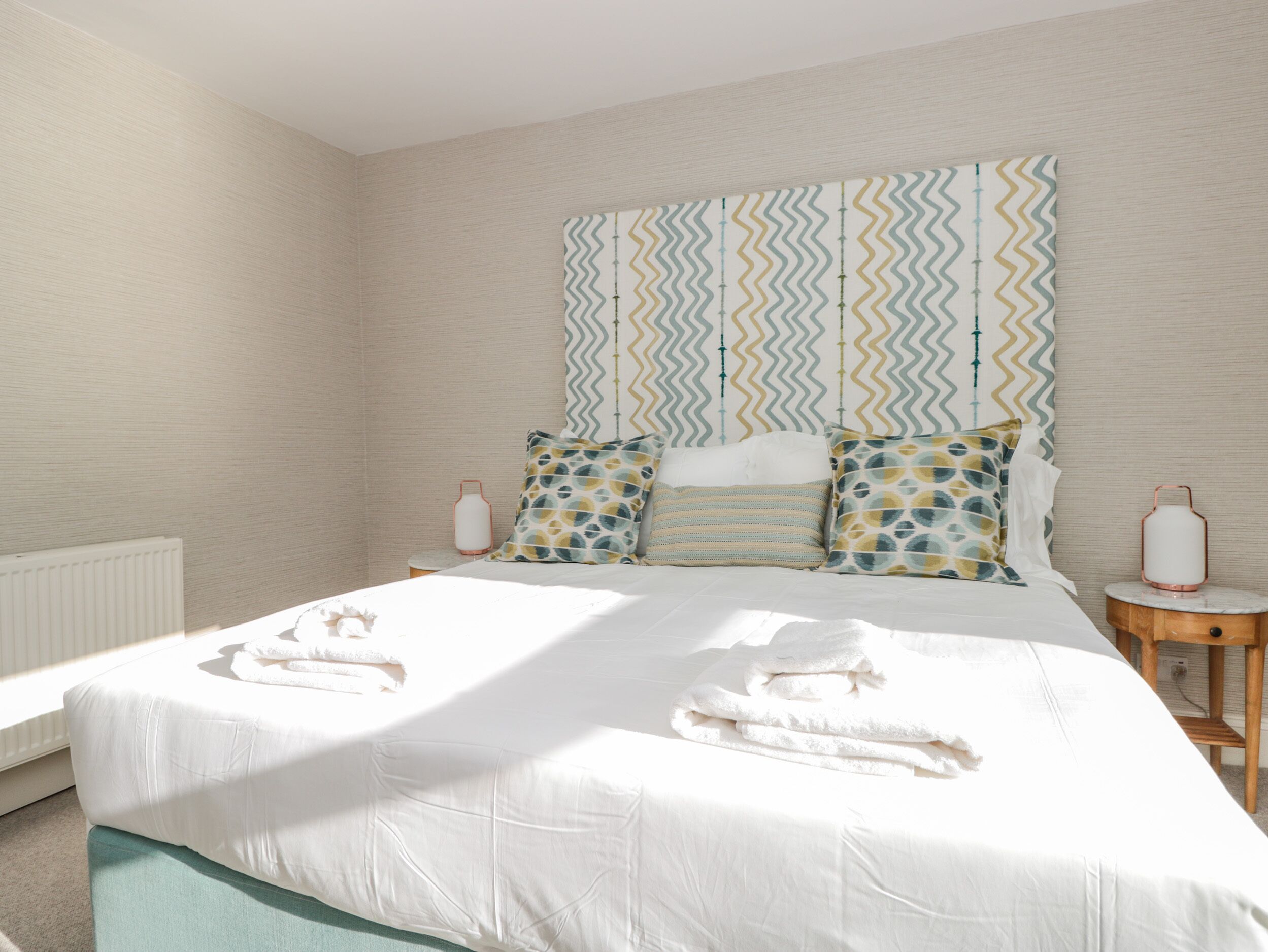 Un letto rifatto con lenzuola e asciugamani bianchi, una parete decorata con un motivo e un comodino con una lampada.