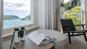 Et værelse med udsigt over en bugt med både, en stol, et bord med kikkert og et magasin, samt en balkon med gelænder.