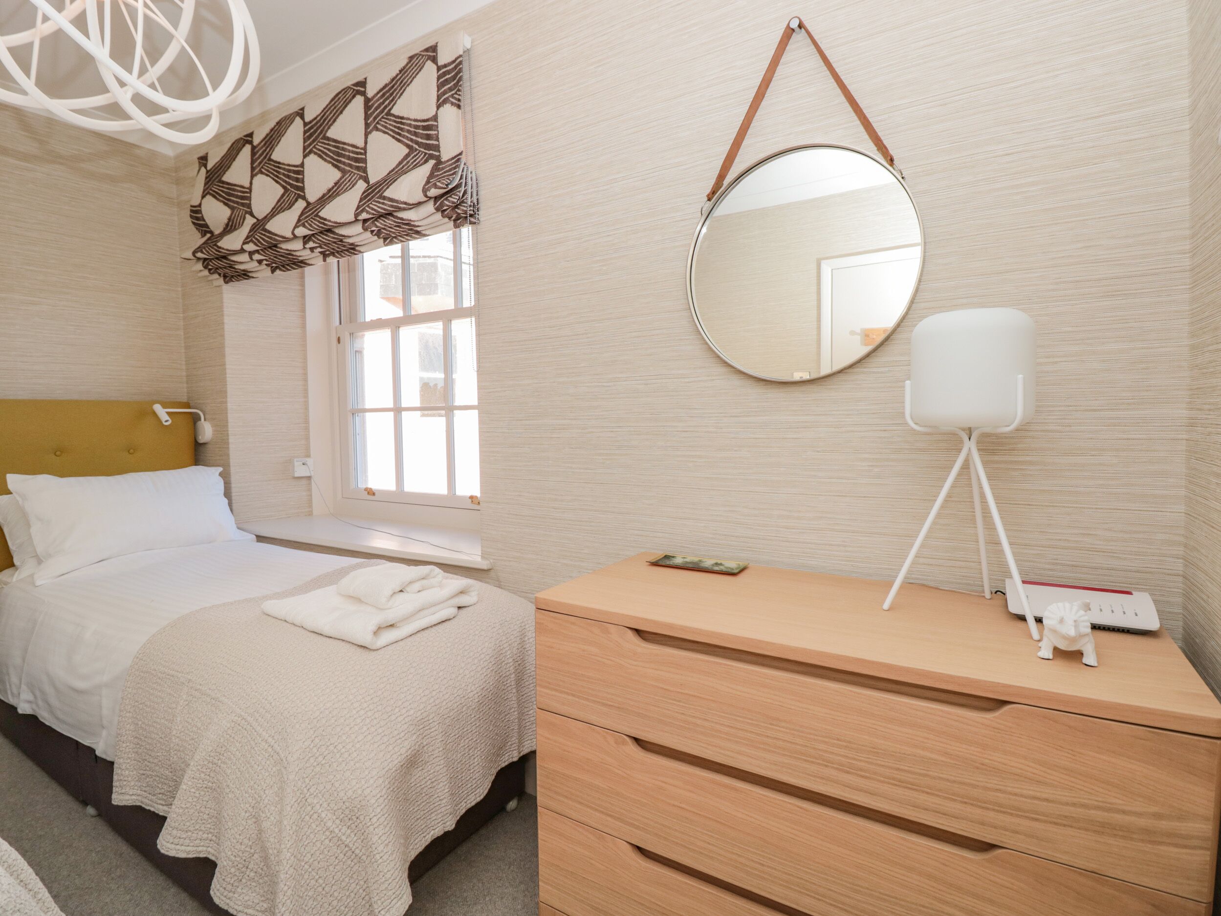 Una camera da letto ordinata con un letto, un comodino con lampada, uno specchio e una finestra con tende a motivi.