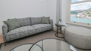 En moderne stue med en grå sofa med et mønster, et rundt sofabord i glas og udsigt til en kystby gennem et stort vindue.