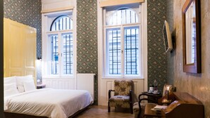 A bedroom with a bed, two chairs, a small table, and a dresser. There are two windows with bars, and a radiator is visible. - Metropolis Budapest Boutique Hotel (Budapest)