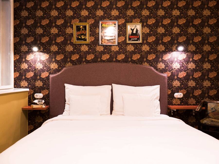 A neatly made bed with two pillows, a wooden nightstand with a lamp, and a chair with a patterned cushion. There are three framed posters on the wall above the bed.