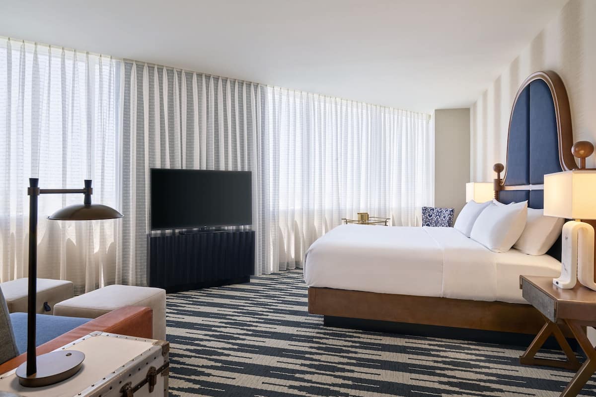A hotel room with a large bed, a flat-screen TV, a desk with a lamp, and a suitcase on the floor.