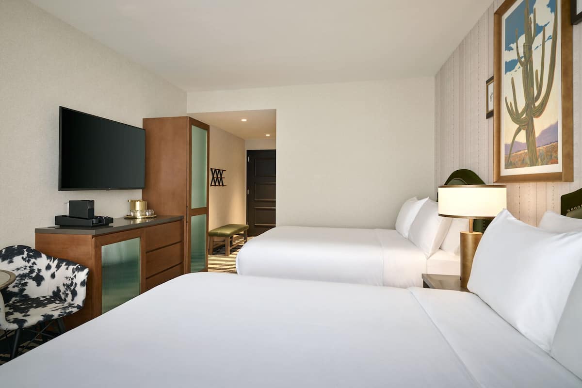 A hotel room with two beds, a desk with a flat-screen TV, a chair, a desk lamp, and a painting on the wall.