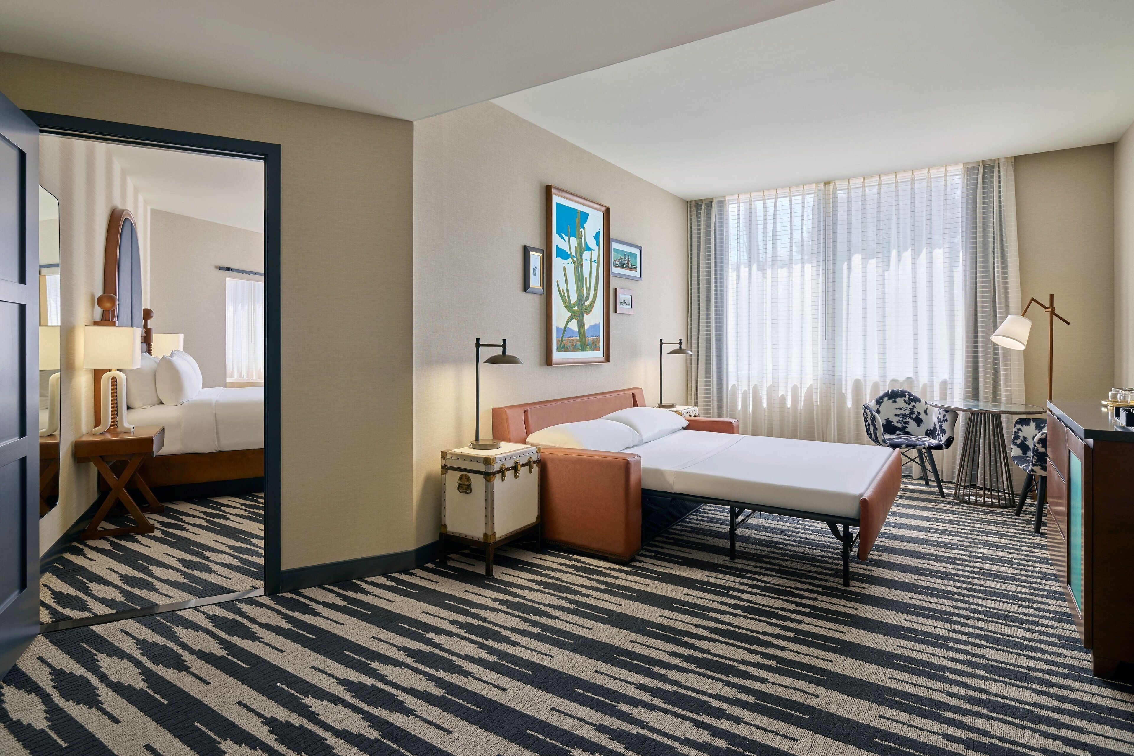Ein Hotelzimmer mit Bett, Nachttischen, Schreibtisch, Stuhl, Lampe, Spiegel und einem Fenster mit Vorhängen.