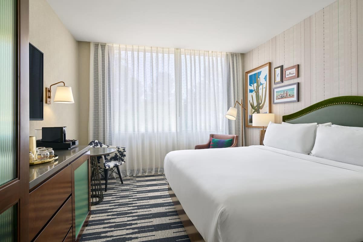 A hotel room with a large bed, a desk with a lamp, a chair, a small table with a tray of items, and a window with sheer curtains.
